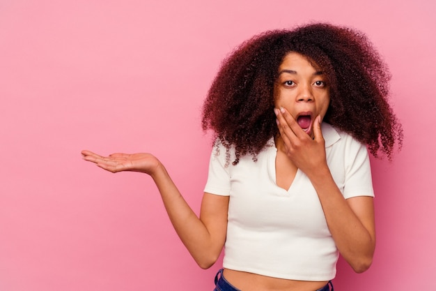 Young african american woman isolated on pink wall holds copy space on a palm, keep hand over cheek. Amazed and delighted.