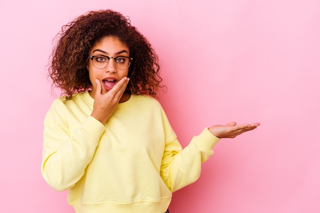 Young african american woman isolated on pink wall holds copy space on a palm, keep hand over cheek. Amazed and delighted.