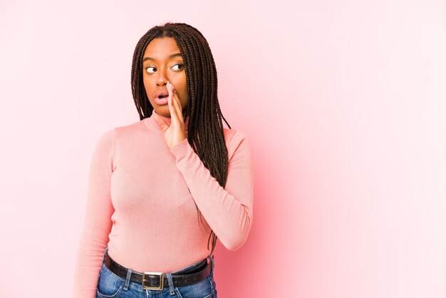 Young african american woman isolated on a pink wall is saying a secret hot braking news and looking aside