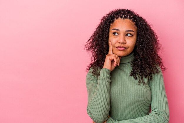 Giovane donna afroamericana isolata sulla parete rosa che osserva obliquamente con espressione dubbiosa e scettica.