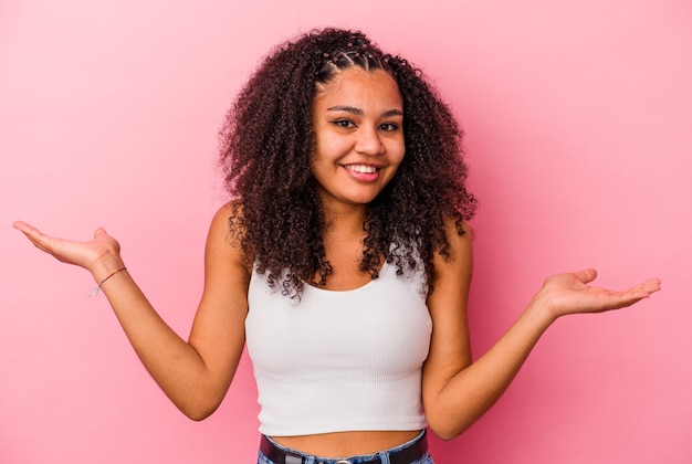 ピンクの壁に隔離された若いアフリカ系アメリカ人の女性は腕でスケールを作り、幸せで自信を持って感じます
