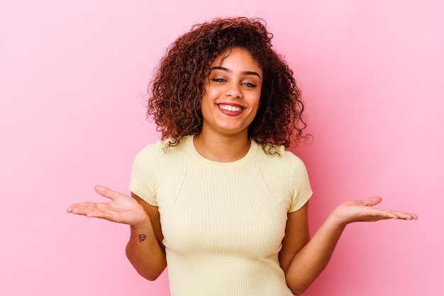 Young african american woman isolated on pink wall makes scale with arms, feels happy and confident.