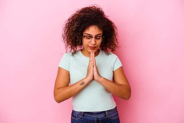 Молодая афро-американская женщина изолирована на розовой стене, молясь, показывая преданность, религиозный человек ищет божественное вдохновение.