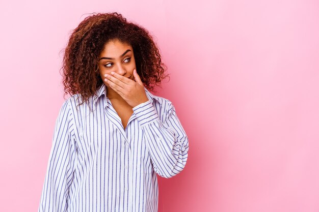 Young african american woman isolated on pink wall thoughtful looking to a copy space covering mouth with hand