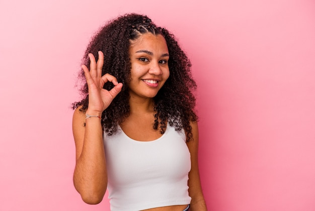 Young african american woman isolated on pink wall winks an eye and holds an okay gesture with hand