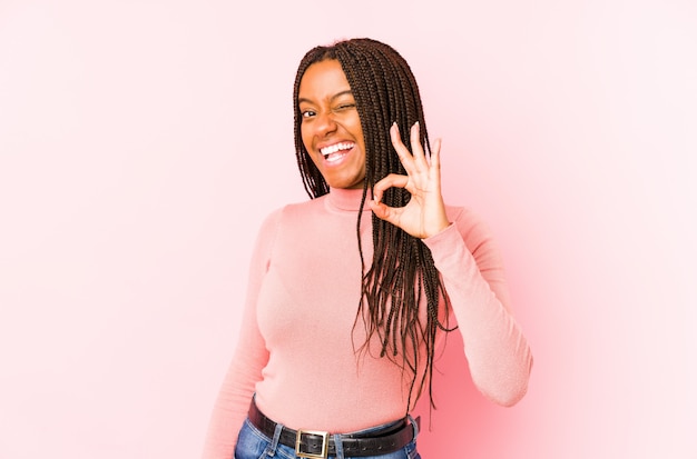 La giovane donna afroamericana isolata su una parete rosa fa l'occhiolino di un occhio e tiene un gesto giusto con la mano.