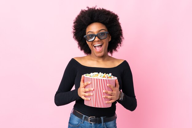 Giovane donna afroamericana isolata sul colore rosa con gli occhiali 3d e che tiene un grande secchio di popcorn