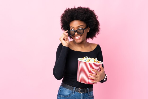 Young African American woman isolated on pink with 3d glasses and holding a big bucket of popcorns