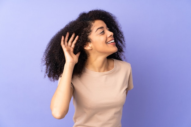 Young african american woman isolated on purple listening to something by putting hand on the ear