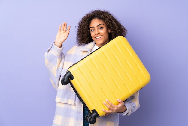 Young African American woman isolated on purple space in vacation with travel suitcase and saluting