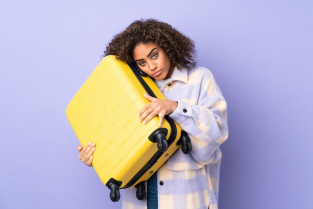 Молодая Афро-американская женщина изолированная на пурпуре в каникулах с чемоданом перемещения и несчастная