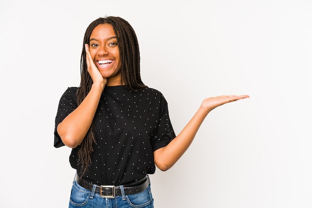 Young african american woman isolated on white wall holds copy space on a palm, keep hand over cheek. Amazed and delighted.