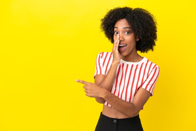 Photo young african american woman isolated on yellow background pointing to the side to present a product and whispering something