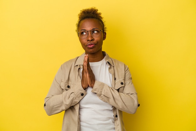 Молодая афро-американская женщина, изолированная на желтом фоне, молясь, показывая преданность, религиозный человек, ищущий божественного вдохновения.