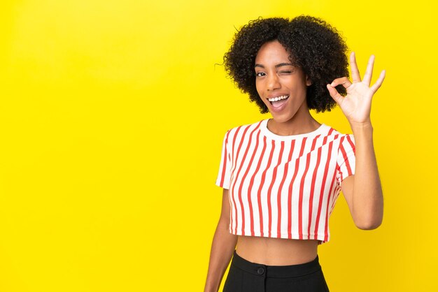 Молодая афро-американская женщина изолирована на желтом фоне, показывая пальцами знак ОК