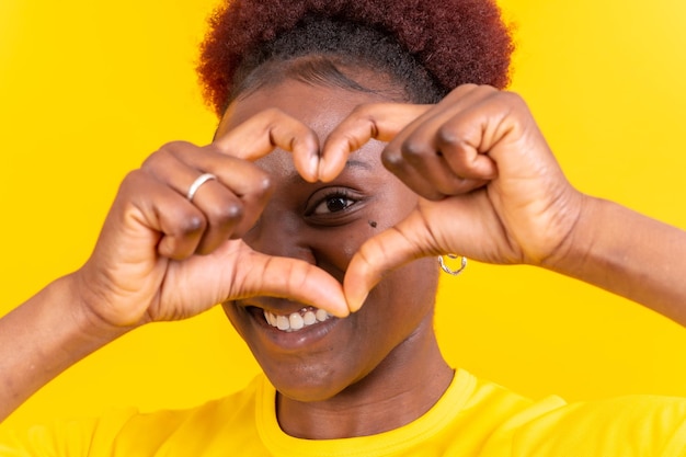 Young african american woman isolated on a yellow background smiling and heart gesture studio shoot