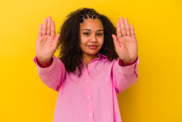黄色い背景に立っている若いアフリカ系アメリカ人女性が、一時停止の標識を示し、あなたを妨げている差し伸べられた手で立っています。
