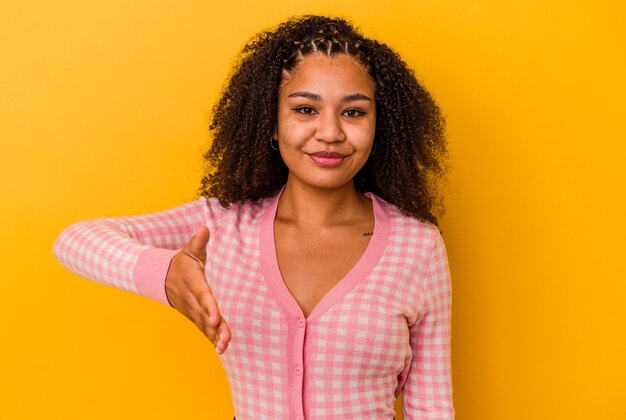 Giovane donna afroamericana isolata su fondo giallo che allunga la mano alla macchina fotografica nel gesto di saluto.