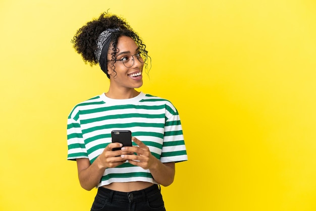 Young african american woman isolated on yellow background using mobile phone and looking up