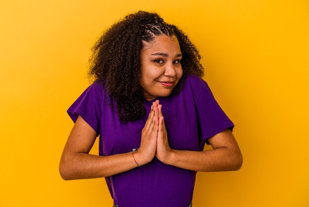 Young african american woman isolated on yellow wall holding hands in pray near mouth, feels confident