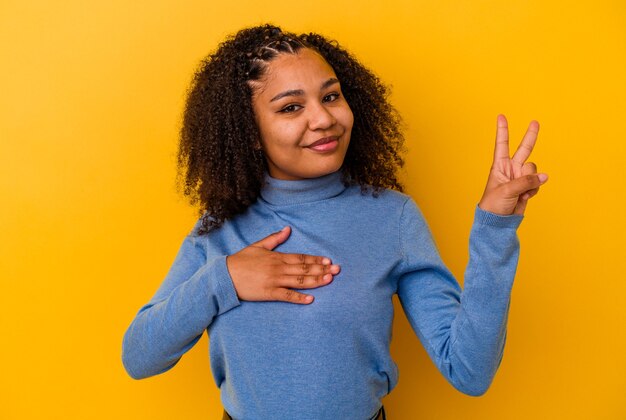 Giovane donna afroamericana isolata sulla parete gialla che presta giuramento, mettendo la mano sul petto.