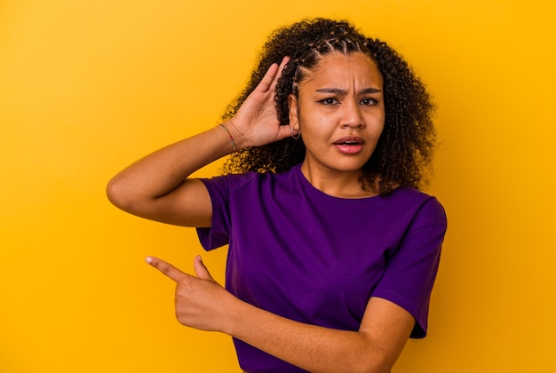 Foto giovane donna afroamericana isolata sulla parete gialla che prova ad ascoltare un pettegolezzo.