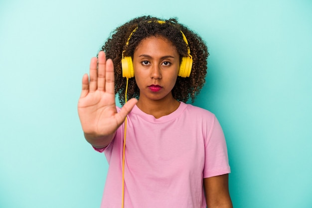 Young african american woman listening to music isolated on blue background standing with outstretched hand showing stop sign, preventing you.