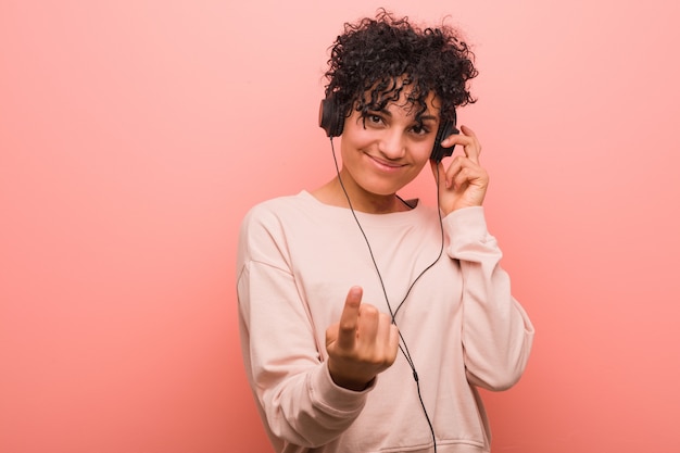 Young african american woman listening to music pointing with finger at you as if inviting come closer.