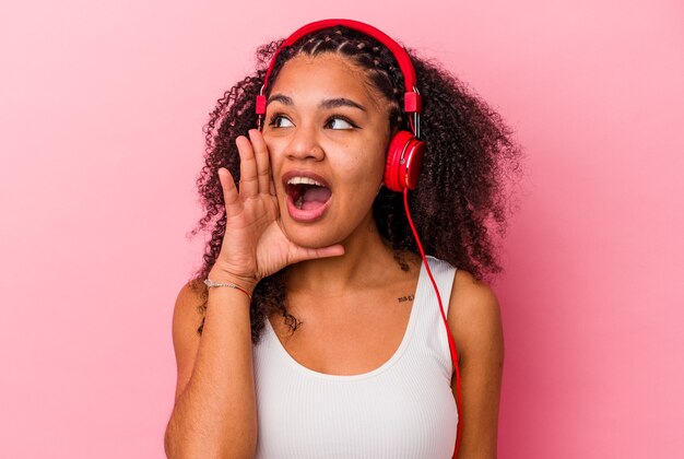 Young african american woman listening to music with headphones isolated on pink background shouting and holding palm near opened mouth.