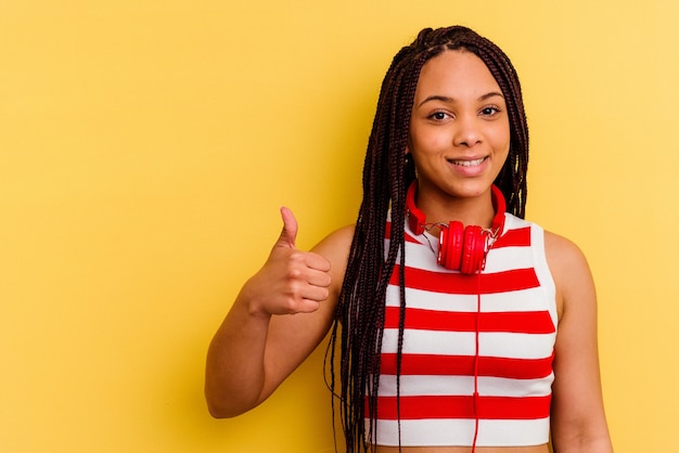 Молодая афро-американская женщина слушает музыку в наушниках, изолированных на желтом фоне, улыбается и поднимает палец вверх