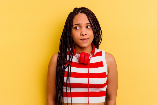 Young african american woman listening to music with headphones isolated on yellow wall confused, feels doubtful and unsure