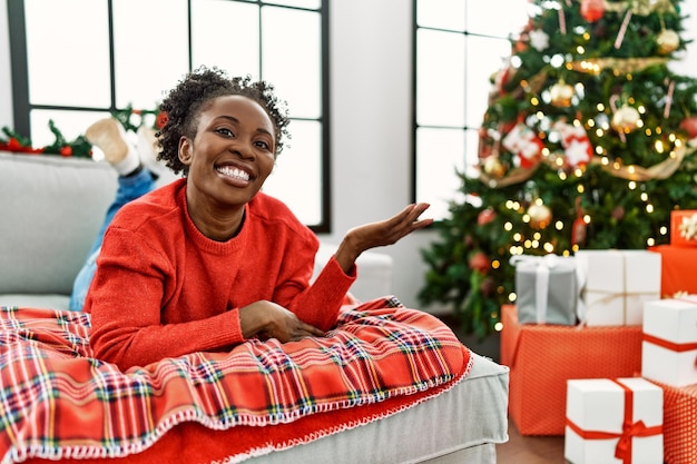 Young african american woman lying on the sofa by christmas tree smiling cheerful presenting and pointing with palm of hand looking at the camera.