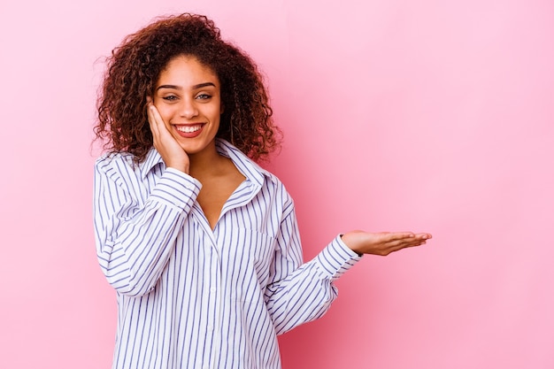 Young african american woman on pink holds copy space on a palm, keep hand over cheek. Amazed and delighted.