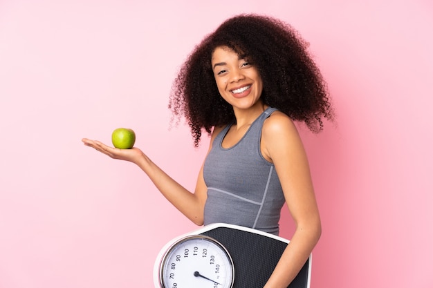 Young african american woman on pink wall with weighing machine and with an apple