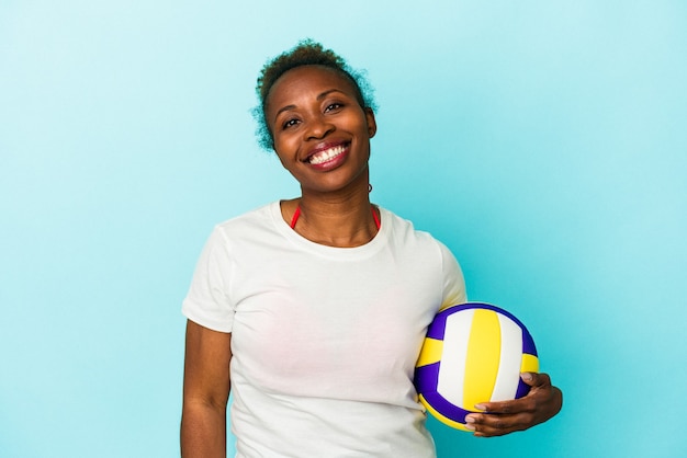 Giovane donna afroamericana che gioca a pallavolo isolata su fondo blu felice, sorridente e allegra.