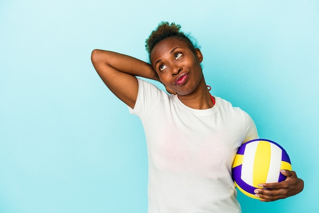 Giovane donna afroamericana che gioca a pallavolo isolata su fondo blu che tocca la parte posteriore della testa, pensa e fa una scelta.