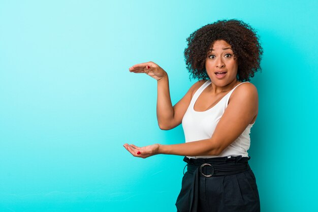 Young african american woman shocked and amazed holding a copy space between hands.