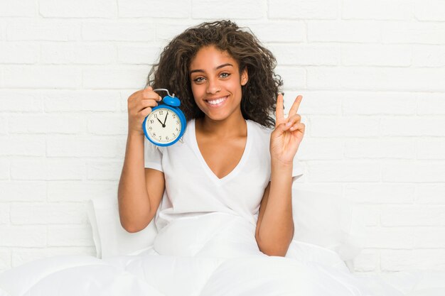 Young african american woman sitting on the bed holding an alarm clock showing number two with fingers.