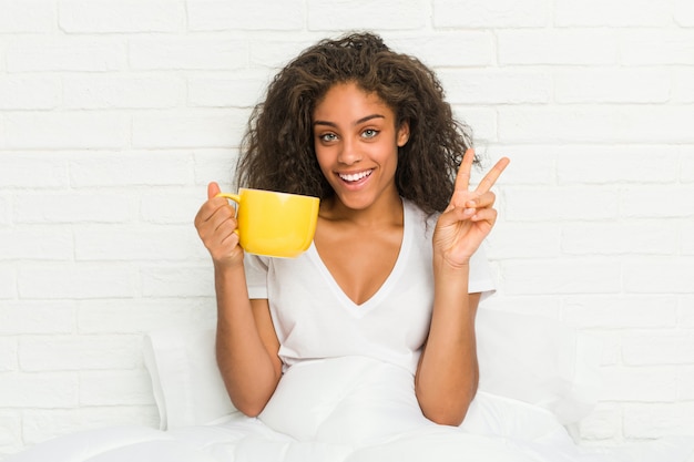 Young african american woman sitting on the bed holding a coffee mug showing number two with fingers.