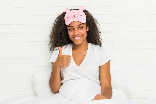 Young african american woman sitting on the bed wearing a sleep mask smiling and raising thumb up