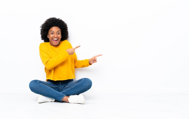 Young African American woman sitting on the floor surprised and pointing side