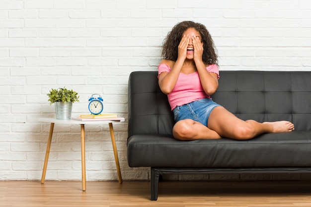 Young african american woman sitting on the sofa covers eyes with hands, smiles broadly waiting for a surprise.