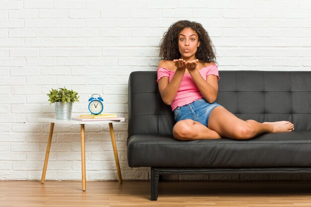 Photo young african american woman sitting on the sofa folding lips and holding palms to send air kiss.