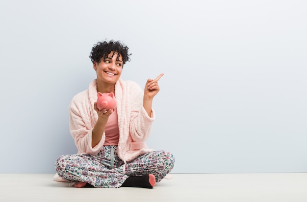 Young african american woman sitting with a piggy bank smiling cheerfully pointing with forefinger away.