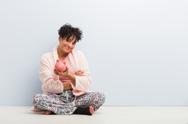 Young african american woman sitting with a piggy bank smiling confident with crossed arms.