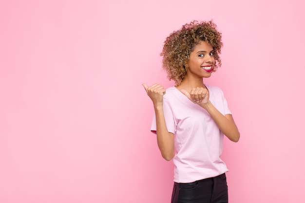 Young african american woman smiling cheerfully and casually pointing to copy space on the side, feeling happy and satisfied against pink wall