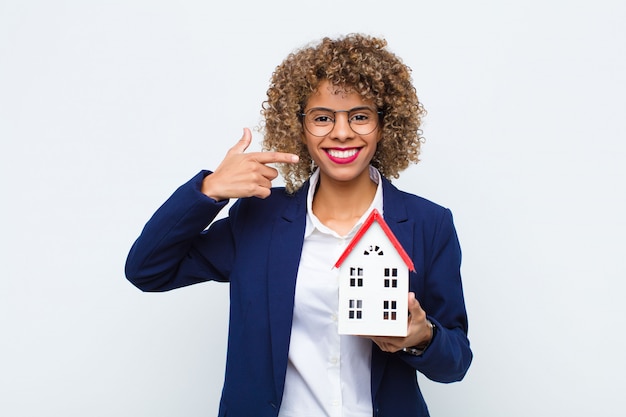 Photo young african american woman smiling confidently pointing to own broad smile, positive, relaxed, satisfied attitude with house model