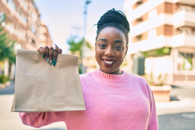 Молодая африканская американка счастливо улыбается, держа бумажный пакет для доставки с едой на вынос в городе