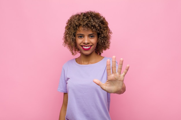 Young african american woman smiling and looking friendly, showing number five or fifth with hand forward, counting down against pink wall