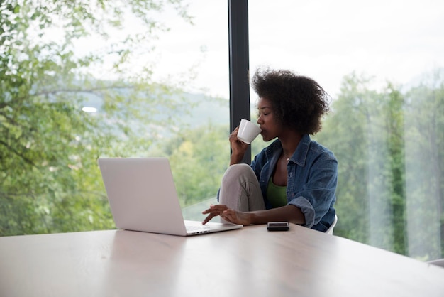 Young african american woman smiling sitting near bright window while looking at open laptop computer on table and holding white mug in her luxury home